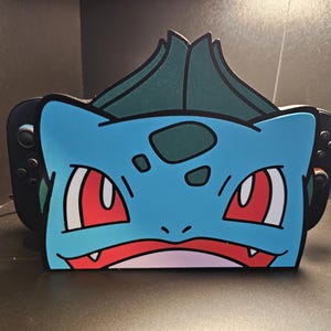 以下が含まれることがあります： 青と緑のフシギダネをテーマにしたNintendo Switchドック。ドックは、赤い目と白い口を持つフシギダネの顔が特徴です。上部には緑の葉のデザインがあります。Switchコントローラーが両側に表示されています。