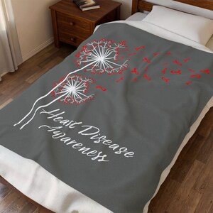 May include: A gray blanket with a dandelion design in white and red, with the words "Heart Disease Awareness" in white script. The blanket is on a bed with a white pillow and a wooden nightstand.