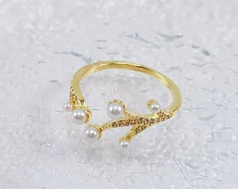 May include: A gold ring with a branch-like design. The ring features small, round, white pearls and clear crystals. The ring is set against a light, textured background.