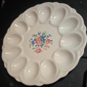 May include: A round white ceramic egg serving platter featuring a scalloped edge and twelve oval-shaped indentations for holding eggs. The center of the platter is decorated with a colorful floral bouquet print featuring pink roses and blue flowers.