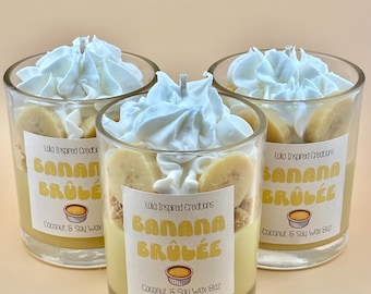 May include: Three glass candles with a banana brûlée theme. Each candle has a yellow wax base, topped with banana slices, whipped cream, and a label that reads "Banana Brûlée". The candles are made with coconut and soy wax.