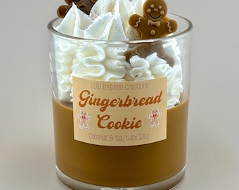 May include: A clear glass candle with a brown wax base, topped with white whipped wax and two gingerbread man decorations. The candle has a label that reads "Gingerbread Cookie" and "Coconut & Soy Wax 12oz."