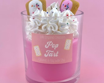 May include: A pink glass candle jar with a white lid. The candle is topped with white frosting and two Pop-Tart pastries. The label reads "Lola Inspired Creations Pop Tart Coconut & Soy Wax 12oz."
