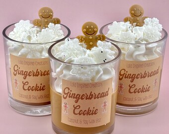 May include: Three clear glass candles with a gingerbread cookie theme. Each candle has a tan wax base, topped with white whipped wax and a gingerbread man. The labels read "Gingerbread Cookie" and "Coconut & Soy Wax 8oz".