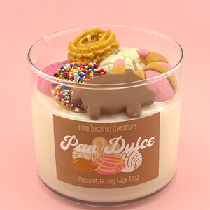May include: A glass jar candle with a brown label that says "Lola Inspired Creations Pan Dulce Coconut & Soy Wax 17oz". The candle is topped with various colorful wax pastries, including a chocolate pig.