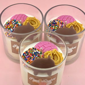 May include: Three glass jars with white soy wax candles decorated with colorful sprinkles, a pink pastry, a brown chocolate piece, and a golden pastry. The jars have labels that say "Lola Inspired Creations" and "Pan Dulce".