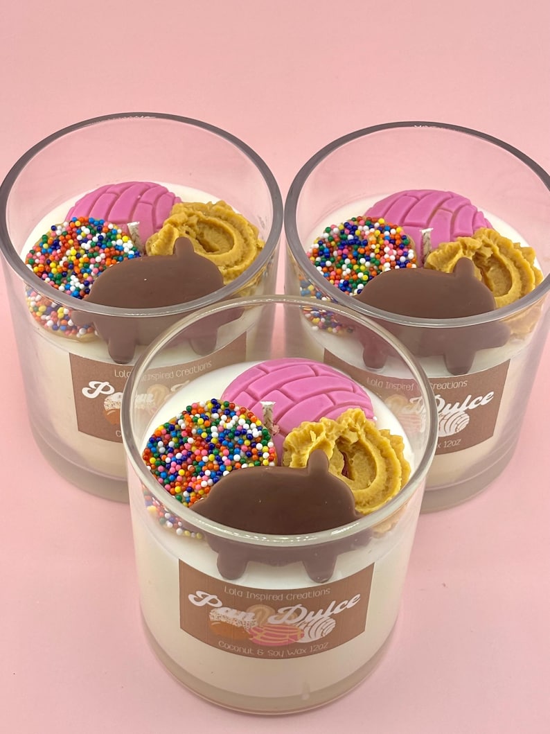 May include: Three glass jars with white soy wax candles decorated with colorful sprinkles, a pink pastry, a brown chocolate piece, and a golden pastry. The jars have labels that say "Lola Inspired Creations" and "Pan Dulce".