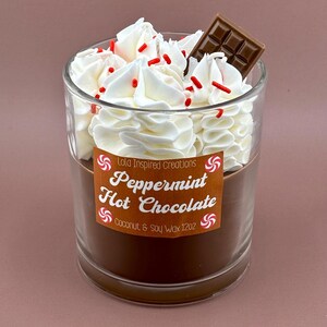 May include: A clear glass candle with a brown base, topped with white whipped wax, red sprinkles, and a chocolate square. The label reads "Peppermint Hot Chocolate" and "Coconut & Soy Wax 12oz."