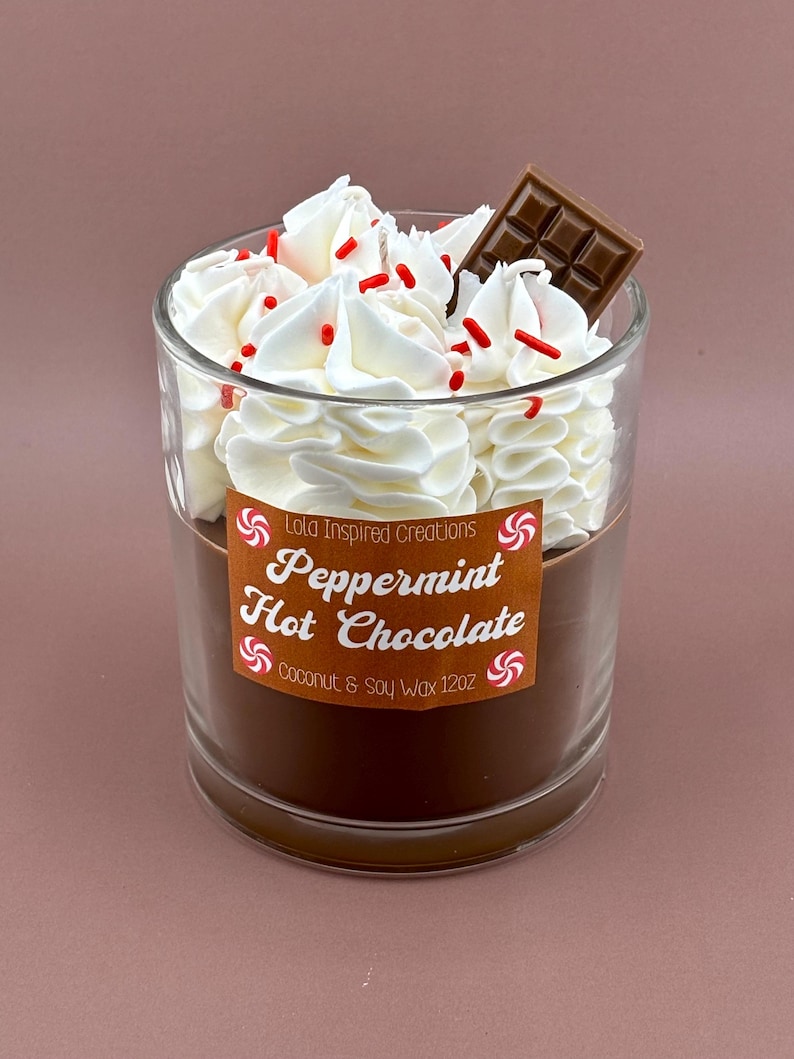May include: A clear glass candle with a brown base, topped with white whipped wax, red sprinkles, and a chocolate square. The label reads "Peppermint Hot Chocolate" and "Coconut & Soy Wax 12oz."