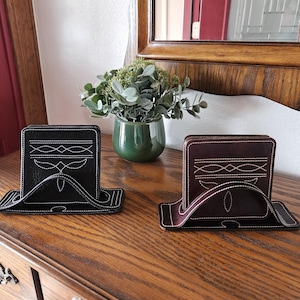 May include: Two leather coasters with matching holders. One is black with white stitching, the other is brown with white stitching. The coasters are on a wooden surface, with a green vase and mirror in the background.