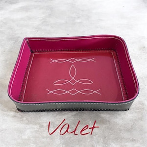 May include: A rectangular valet tray featuring a deep red interior base with white stitched decorative patterns and a vibrant pink inner rim. The exterior is dark gray with black stitched edges, and the word Valet is written below the tray.