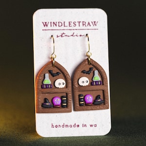 May include: Polymer clay earrings in the shape of bookshelves, with Halloween-themed decorations. Each earring features a bat, skull, potion bottle, books, a witch's hat, and a crystal ball. The earrings are suspended from gold-coloured hooks.