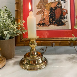 May include: A gold candle holder with a white candle lit inside. The candle holder is on a white surface in front of a framed picture with a red mat.