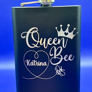 May include: Black stainless steel hip flask with a silver lid and a silver inscription that reads "Queen Bee Katrina" with a crown, heart, and bee graphic.