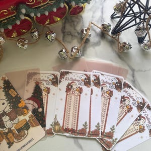 May include: A collection of rectangular paper budgeting challenge cards featuring Snoopy illustrations, holiday themes, and specific dollar amounts like $2, $3, $5, $6, and $7. Each card includes a grid for tracking savings and the text BUDGETINGMYDEBTS.
