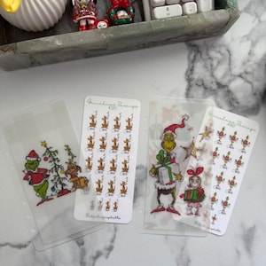 May include: Clear plastic budget inserts with Christmas designs. One insert shows the Grinch with a Christmas tree, another has reindeer, and a third depicts the Grinch with a gift. A calculator and decorative figurines are in the background.