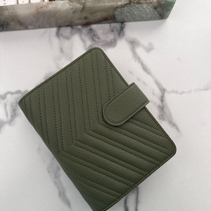 May include: An olive green leather planner cover featuring a textured pebble grain finish and a chevron-patterned quilted stitch design. The rectangular organizer includes a secure tab closure on the right side to keep the contents protected.