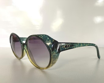 May include: Vintage oversized sunglasses with a green and gold speckled frame and purple gradient lenses. The sunglasses have a rounded shape and a gold logo on the temple. A stylish accessory for eye protection.