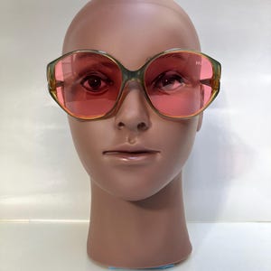 May include: Vintage oversized sunglasses with a green and gold frame and pink lenses. The sunglasses are displayed on a mannequin head. The lenses have the text "HL" printed on them.