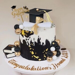May include: A graduation cake with black and white frosting, gold accents, and a mortarboard topper. The cake is decorated with gold and black balloons, a diploma, and the words "Happy Graduation" and "Congratulations Hamad 2021".