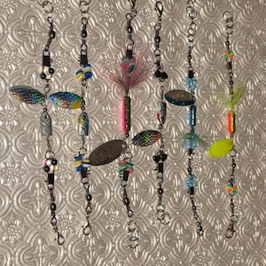 Puede incluir: Una colección de señuelos de pesca con diversos diseños y colores. Cada señuelo presenta un anzuelo metálico, un giratorio y elementos decorativos como palas iridiscentes, plumas coloridas y cuentas. Los señuelos están dispuestos verticalmente sobre un fondo texturizado.