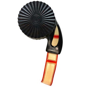 May include: A black plastic tape dispenser featuring a circular, fluted housing and a curved handle. A strip of cream-colored adhesive tape with thin red border lines extends from the dispenser, showing a repeating rectangular pattern.