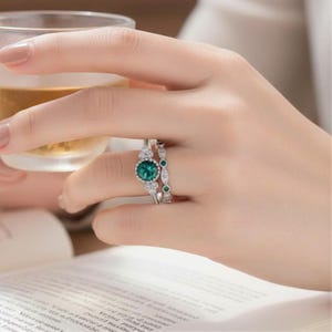 May include: A silver-toned ring set featuring a central round-cut green gemstone surrounded by small clear stones, paired with a matching band containing smaller green stones and clear accents, worn on a finger near a glass of liquid.