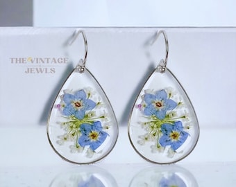 May include: Teardrop-shaped earrings featuring pressed blue forget-me-not flowers and small white blossoms encased in clear resin with silver-toned metal rims and hooks. The text THE VINTAGE JEWELS is printed in black on the upper left side.