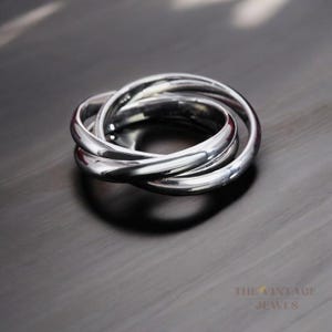 Könnte beinhalten: Ein silberner Ring mit drei ineinandergreifenden Bändern. Der Ring ist glatt und poliert.