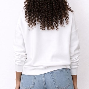 Back view of woman wearing a white Sweatshirt and distressed blue jeans on a plain background.