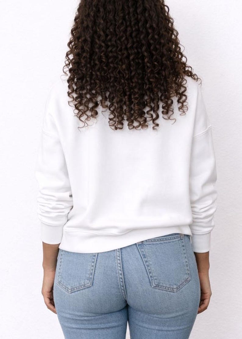 Back view of woman wearing a white Sweatshirt and distressed blue jeans on a plain background.