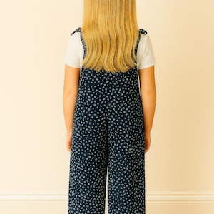 Back view of young girl wearing navy blue patterned Tie Shoulder Jumpsuit with a white shirt underneath, standing against a beige wall.