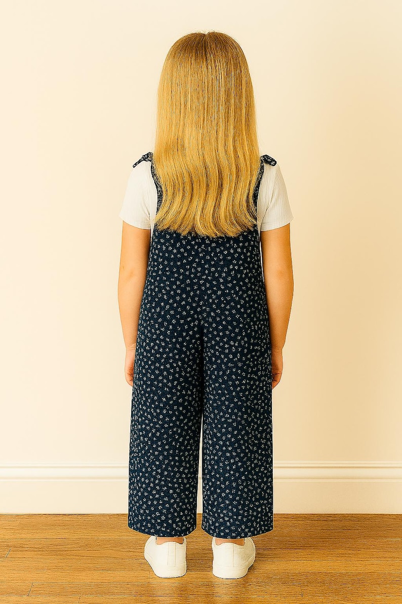 Back view of young girl wearing navy blue patterned Tie Shoulder Jumpsuit with a white shirt underneath, standing against a beige wall.