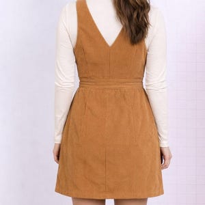 Back view of woman wearing a brown corduroy Button Down Overall Dress with a white turtleneck and brown boots on a plain background.