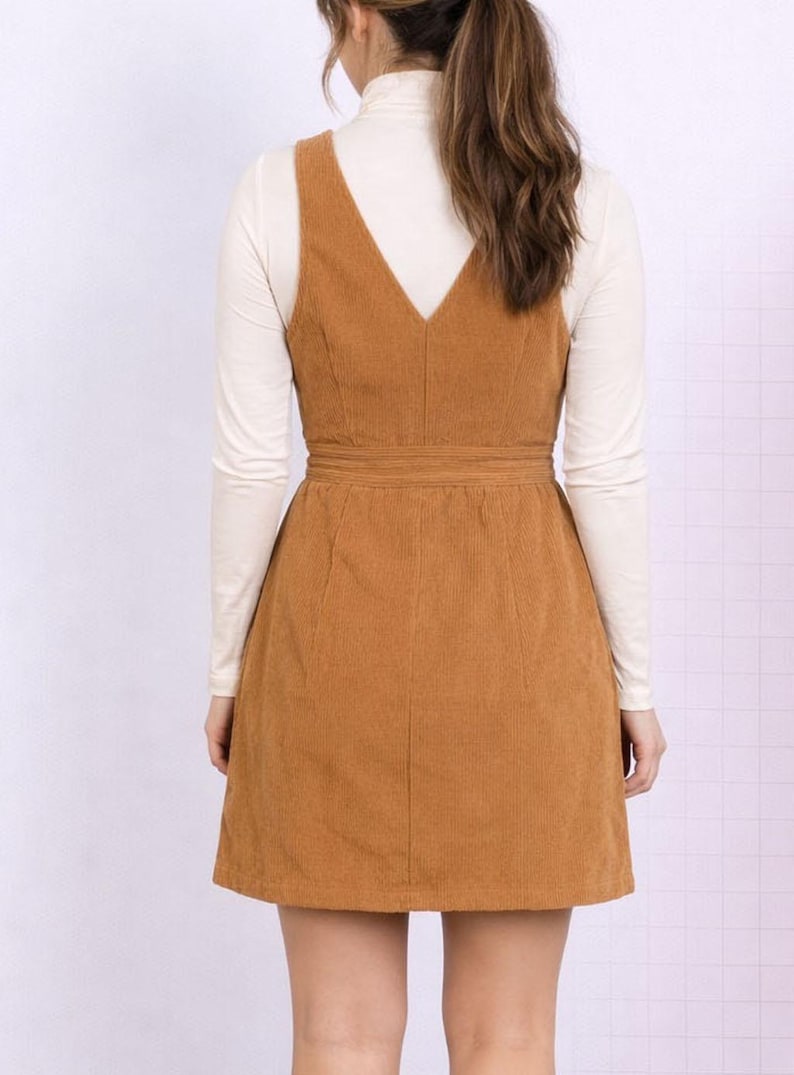 Back view of woman wearing a brown corduroy Button Down Overall Dress with a white turtleneck and brown boots on a plain background.