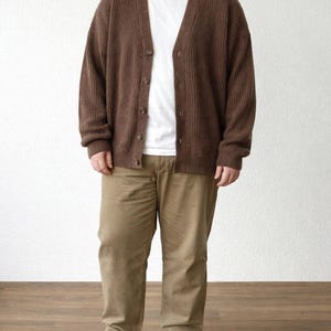Person wearing a brown Plus Size Button Front Cardigan, white shirt, black pants, and blue sneakers against a light pink wall.