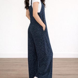 Side view of woman wearing navy Tie Shoulder Jumpsuit with white polka dots and white tee underneath on plain white background.