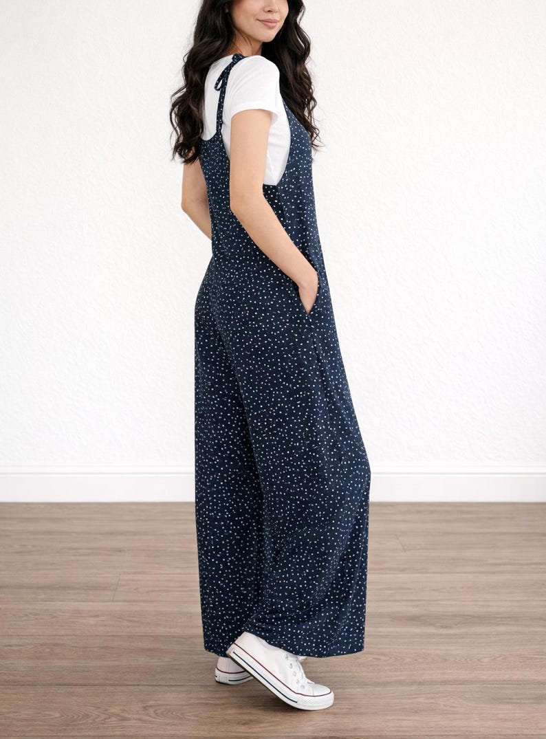 Side view of woman wearing navy Tie Shoulder Jumpsuit with white polka dots and white tee underneath on plain white background.