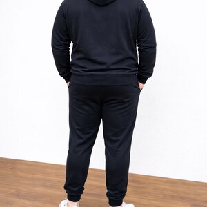 Back view of person wearing a black Plus Size Mens Classic Hoodie and pants with white sneakers on a wooden floor.
