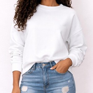 Woman wearing a white Sweatshirt and distressed blue jeans on a plain background.