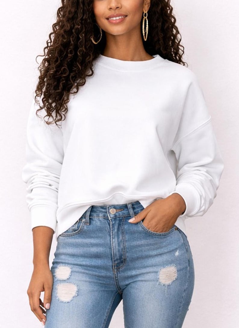 Woman wearing a white Sweatshirt and distressed blue jeans on a plain background.