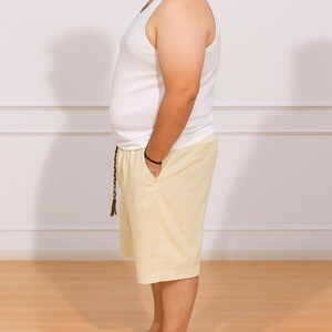 Side view of person wearing a white tank top and beige Plus Size Drawstring Knee Length Shorts standing against a plain white wall.