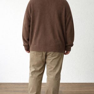 Back view of person wearing a brown Sweater and beige pants standing against a white wall.
