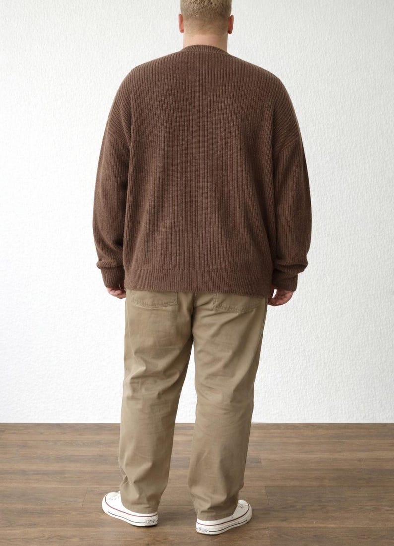Back view of person wearing a brown Sweater and beige pants standing against a white wall.