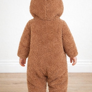 Back view of child wearing a brown fleece onesie with bear ears against a white wall.