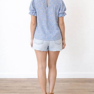 Back view of woman wearing a blue floral Puff Sleeve Blouse and denim shorts on a white background.