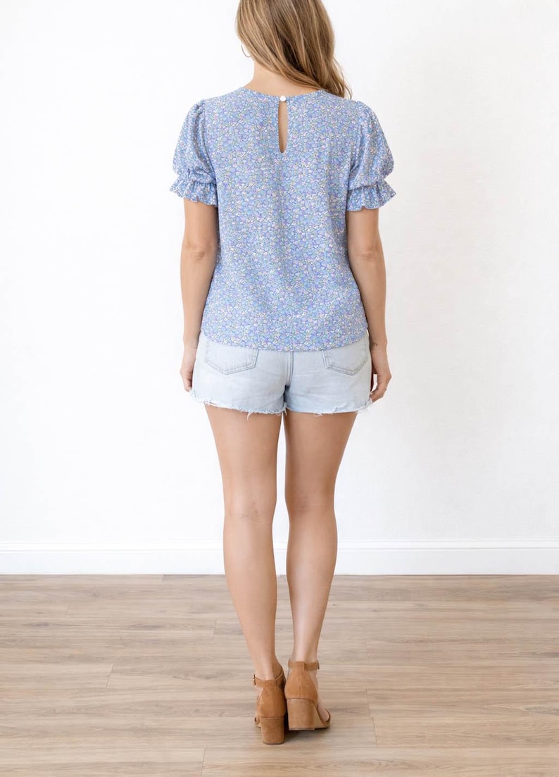 Back view of woman wearing a blue floral Puff Sleeve Blouse and denim shorts on a white background.