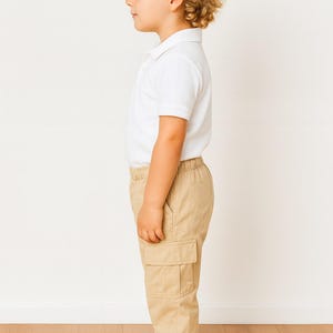 Side view of child wearing a white polo shirt and beige Cargo Pants standing on a wooden floor with a white wall background.