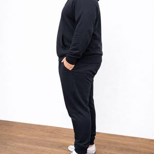 Side view of person wearing a black Plus Size Mens Classic Hoodie and pants with white sneakers on a wooden floor.