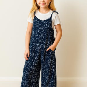 Young girl wearing a navy blue polka dot Tie Shoulder Jumpsuit with a white shirt underneath, standing against a plain wall.